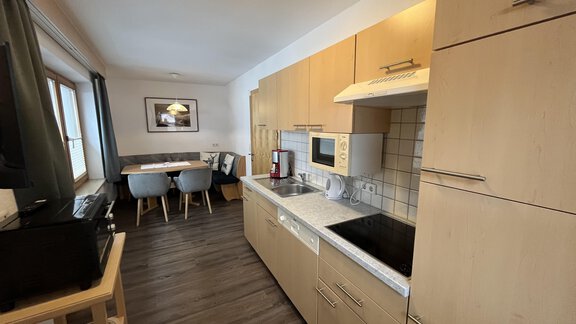 A modern kitchen with wood-colored cabinets and a bright countertop. In the background, there is a dining table with chairs. - Franzele Appartements & Zimmer - Sölden