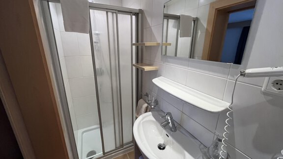 A modern bathroom with a shower, sink, and mirror. The walls are tiled and the furnishings are functional. - Franzele Appartements & Zimmer - Sölden