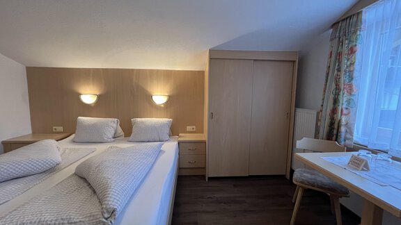 A cozy hotel room with two beds, light walls, and wood paneling. There is a table with chairs and a large window with curtains. - Franzele Appartements & Zimmer - Sölden