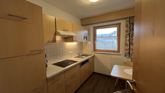 A modern kitchen with wooden-colored cabinets and a window that lets in light. There is a small dining area with a table and a chair. - Franzele Appartements & Zimmer - Sölden