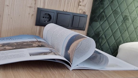 An open magazine lies on a wooden table. In the background, an electrical outlet and a green, padded wall are visible. - Garni Rustica - Sölden