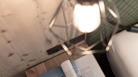 A bedside lamp shines over an open magazine on a wooden table. In the background, part of a bed and a green wall can be seen. - Garni Rustica - Sölden