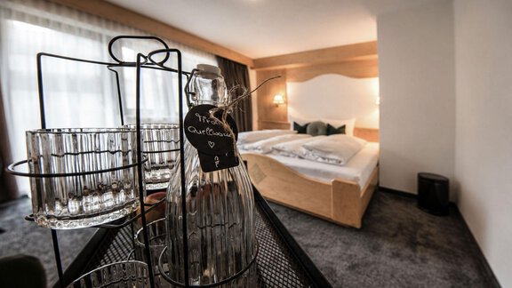 A modern hotel room with a large bed and soft colors. In the foreground, there are glasses and a water bottle on a table. - Garni Rustica - Sölden