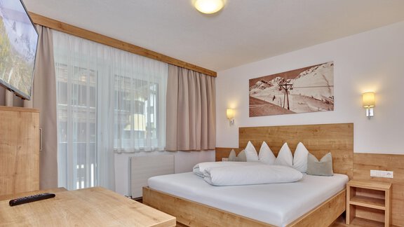 A modern bedroom with a wooden bed and a television. The walls are light, and there are large windows with curtains. - Gästehaus Bergkristall - Sölden