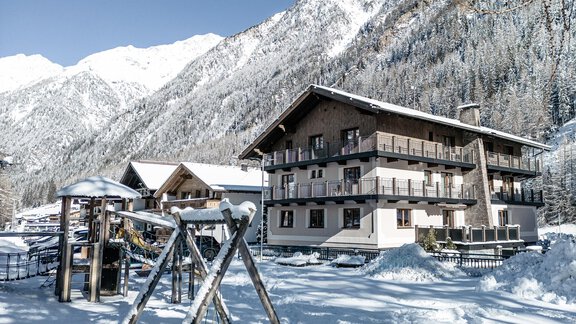 A snow-covered mountain village with a modern building and a playground in the foreground. The surroundings are characterized by impressive mountains and a clear winter landscape. - Gästehaus Dr. Riml - Sölden