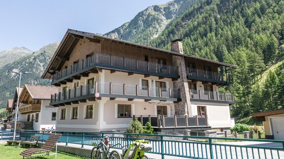 A modern building in alpine style, surrounded by green mountains. In the foreground, there are bicycles and a lounge chair on a lawn. - Gästehaus Dr. Riml - Sölden