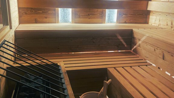 A modern wooden sauna with a wooden bench and a water bucket. Soft light falls through the windows, creating a relaxing atmosphere. - Gästehaus Europa - Sölden
