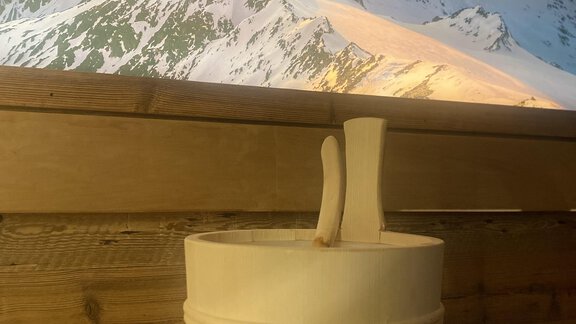 A wooden bucket stands on a wooden bench overlooking snow-covered mountains. In the background, the sun shines over the mountain landscape. - Gästehaus Europa - Sölden
