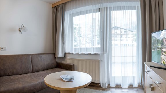 A cozy living room with a brown sofa and a round table. Large windows with curtains provide plenty of daylight. - Gästehaus Europa - Sölden