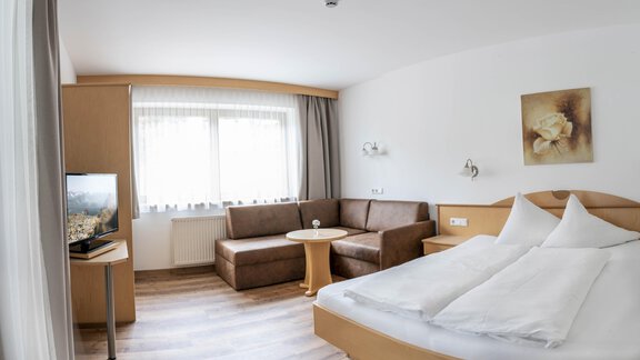 A cozy hotel room with a double bed, a seating area, and a table. Large windows provide plenty of daylight. - Gästehaus Europa - Sölden