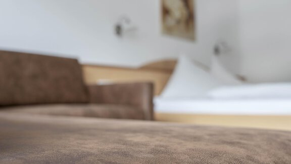 A modern room with a brown sofa in the foreground. In the background, there is a bed and walls in light colors. - Gästehaus Europa - Sölden