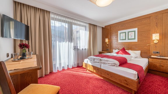 A cozy hotel room with wood paneling and a red carpet. There is a large window with curtains and a TV on the wall. - Gästehaus Maier - Sölden