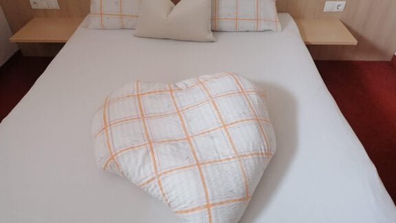 A cozy bed with white pillows and a heart-shaped blanket in the middle. The room has a warm atmosphere with red carpet flooring. - Gästehaus Sternberger - Sölden