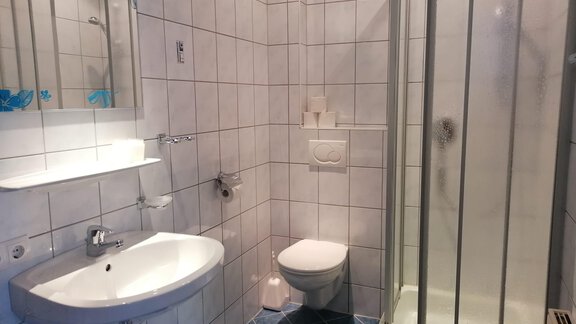 A modern bathroom with white tiles and blue accents. It includes a shower, a sink, and a toilet. - Gästehaus Sternberger - Sölden