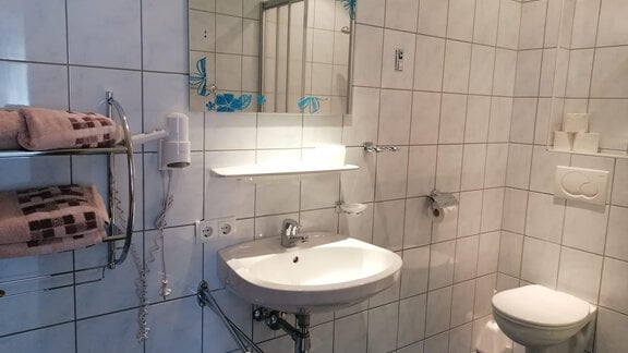 A modern bathroom with white tiles and a large mirror. There is a sink, a toilet, and towels on a holder. - Gästehaus Sternberger - Sölden