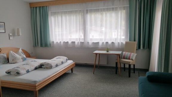 A simple and bright room with a bed, a table, and a chair. The large windows let in plenty of light and the curtains are in soft colors. - Gästehaus Sternberger - Sölden