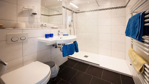 A modern bathroom with a shower and white tiled walls. There is a sink area with blue towels and an elegant decor. - Gästehaus Veit Fiegl - Sölden