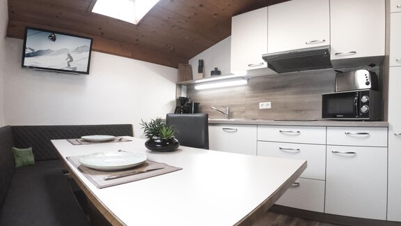 A modern kitchen with a white table and gray chairs. A television and a plant arrangement complement the cozy atmosphere. - Gästeheim Schmiedhof - Sölden