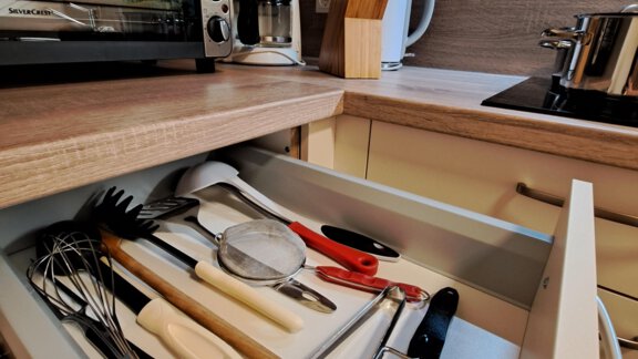 A modern kitchen with an open drawer full of cooking utensils. In the background, kitchen appliances like an oven and a coffee maker are visible. - Gästeheim Schmiedhof - Sölden
