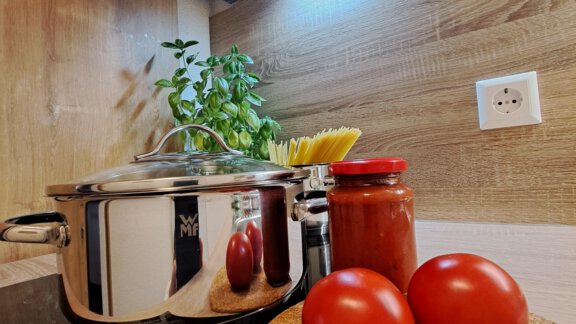 A modern kitchen with a cooking pot, fresh tomatoes, and a jar of tomato sauce. On the wooden board, there is also a small pot with fresh herbs. - Gästeheim Schmiedhof - Sölden