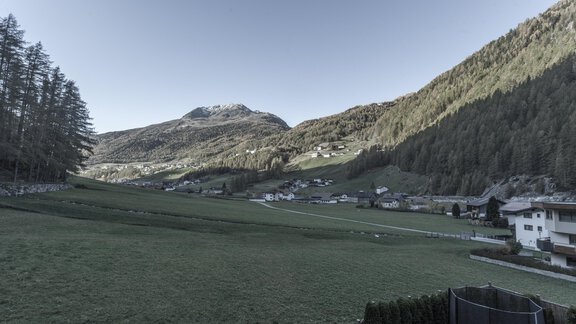 A tranquil mountain landscape with gentle hills and scattered houses. In the background, forested mountains and a clear sky can be seen. - Gästeheim Schmiedhof - Sölden