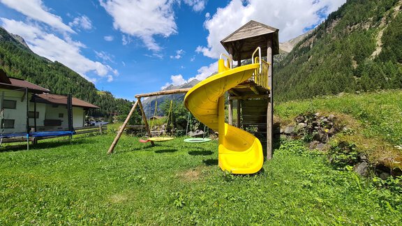 A playground with a yellow slide and swings. In the background, mountains and a blue sky are visible. - Gästeheim Schmiedhof - Sölden