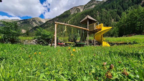A playground with a slide and swings is situated on a green meadow. In the background, mountains and a blue sky can be seen. - Gästeheim Schmiedhof - Sölden