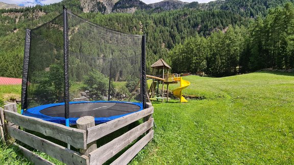 A trampoline and a slide are on a green meadow, surrounded by mountains and trees. The sky is blue with some clouds. - Gästeheim Schmiedhof - Sölden