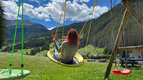 A swinging person with red hair is sitting on a swing in a green meadow. In the background, there are mountains and a blue sky with a few clouds. - Gästeheim Schmiedhof - Sölden