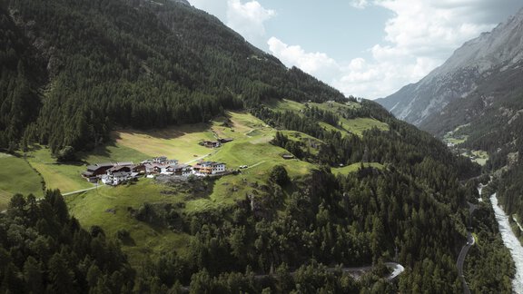 A picturesque landscape with green hills and a small village. In the background, high mountains and a clear sky can be seen. - Gasthof Granstein - Sölden