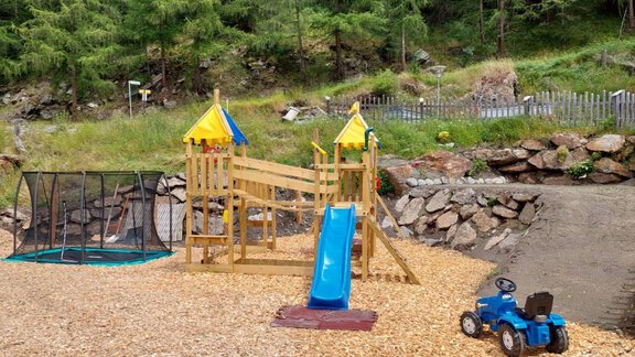 A playground with a wooden structure, slide and a small tractor. The area is covered with wood chips and surrounded by trees. - Granbichlhof - Sölden