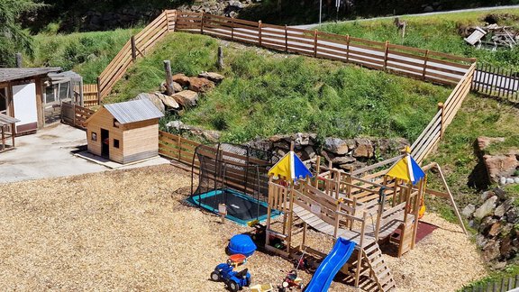 A playground with a large wooden play structure, slide, and colorful flags. In the background, a garden house and a play area with toys can be seen. - Granbichlhof - Sölden