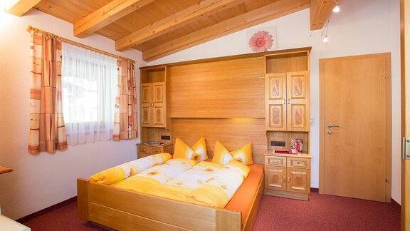 A cozy bedroom with a wooden ceiling and a large bed in warm colors. There are wooden wardrobes and a window with curtains that let in natural light. - Haus Alois - Sölden