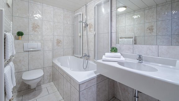 A modern bathroom with tiled walls and a large mirror. There is a bathtub, a shower, and a sink, as well as towels and a small plant for decoration. - Haus Amaris - Sölden