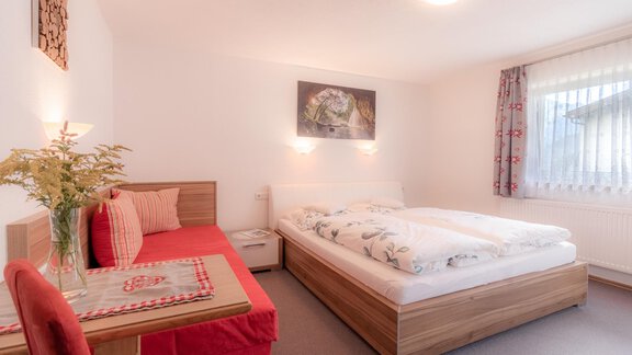 A cozy bedroom with a large bed and a red sofa. It has bright walls and plenty of daylight through the window. - Haus Edmund - Sölden