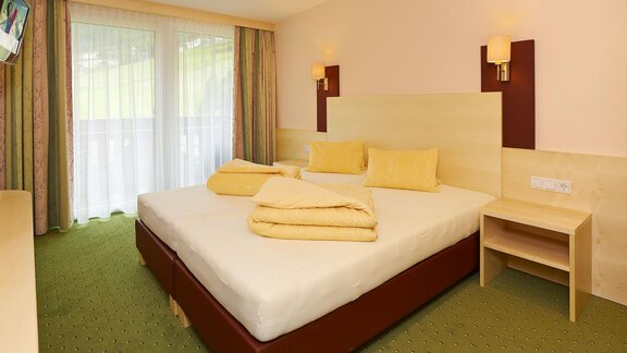 A bright bedroom with a large double bed and cozy yellow cushions. The room has large windows and a green carpet. - Haus Fridolin - Sölden