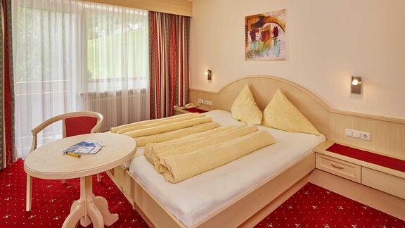 A cozy hotel room with a double bed, two large yellow pillows, and a small table. Large windows provide plenty of daylight and a pleasant atmosphere. - Haus Fridolin - Sölden