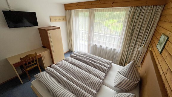 A cozy bedroom with a double bed and striped pillows. The room has large windows with curtains and a desk in the corner. - Haus Nesselhof - Sölden