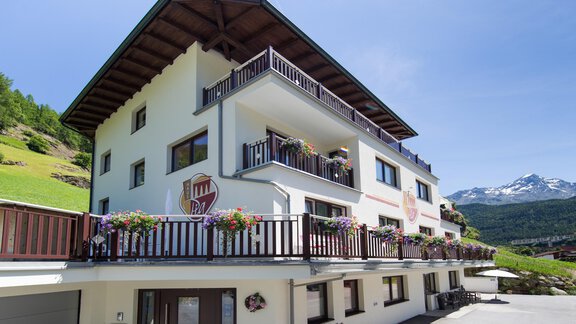 A modern building with several balconies and many flower boxes. In the background, green hills and snow-covered mountains can be seen. - Haus PIA by A CASA - Sölden
