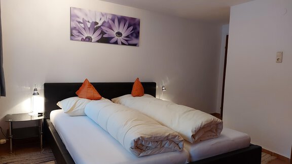 A modern bedroom with a large bed and orange pillows. A picture of purple flowers hangs on the wall. - Haus Pitzwald - Sölden
