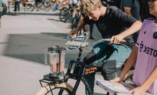 A boy rides a special bicycle that powers a blender. In the background, additional bicycles and participants at an event can be seen. - Haus Rauch - Sölden