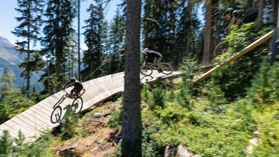 A cyclist rides on a wooden trail through a forest. Surrounded by tall trees and a sunny mountain landscape. - Haus Rauch - Sölden