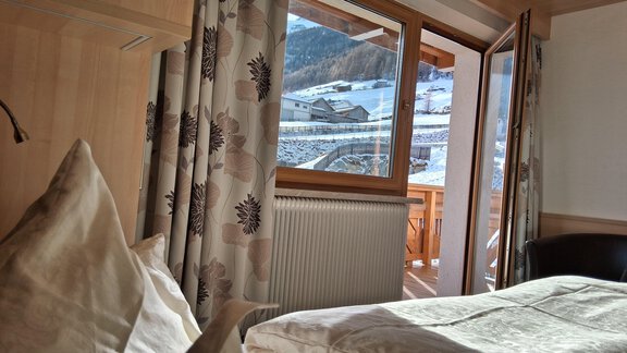 A cozy room with a bed and large windows overlooking a snowy landscape. The curtains and wooden elements give the space a warm atmosphere. - Haus Rehwinkl - Sölden