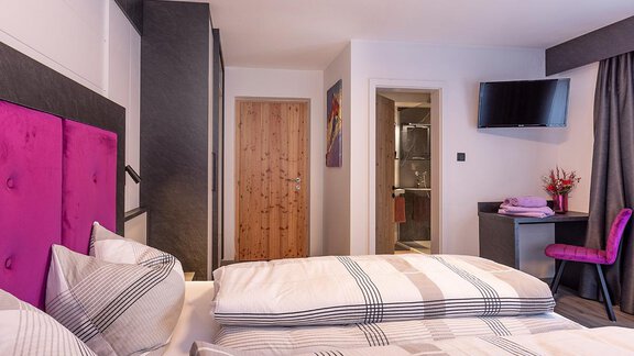 A modern hotel room with two beds and a purple headboard. There is a television on the wall and a table with a chair. - Haus Sieglinde Color - Sölden