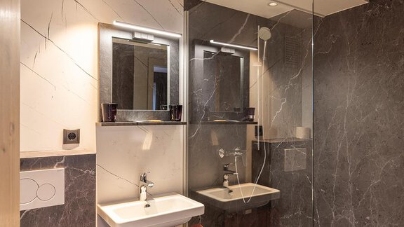 A modern bathroom with dark gray marble walls. There is a sink, a large mirror, and a shower. - Haus Sieglinde Color - Sölden