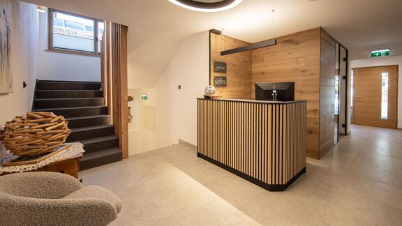 A modern reception area with a wooden desk and a staircase in the background. The walls are bright and the lighting is friendly. - Haus Sonnwend - Sölden