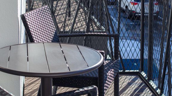 A small balcony with a round table and two chairs. In the background, cars and a modern building are visible. - Haus Sonnwend - Sölden