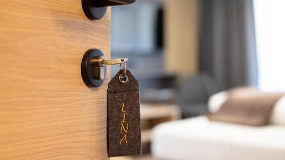 A room key named "LINA" hangs on the door of a hotel room. In the background, a modern and stylish room can be seen. - Haus Sonnwend - Sölden