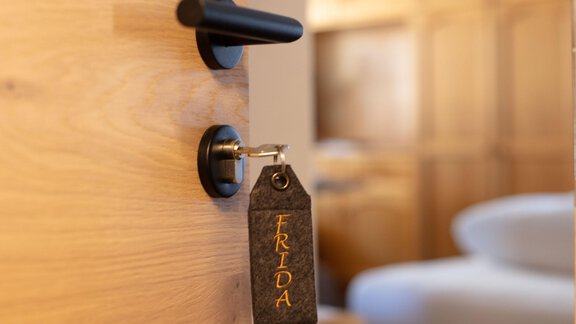 A room key named "FRIDA" hangs on a door. The door has a modern door handle and you can see a blurred background of a cozy room. - Haus Sonnwend - Sölden