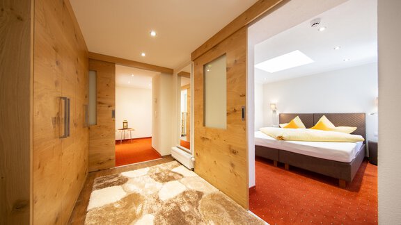 A cozy hallway with wooden paneling and carpet leads into a bright bedroom. The bedroom features a double bed with yellow pillows and plenty of natural light. - Haus Stefanie - Sölden